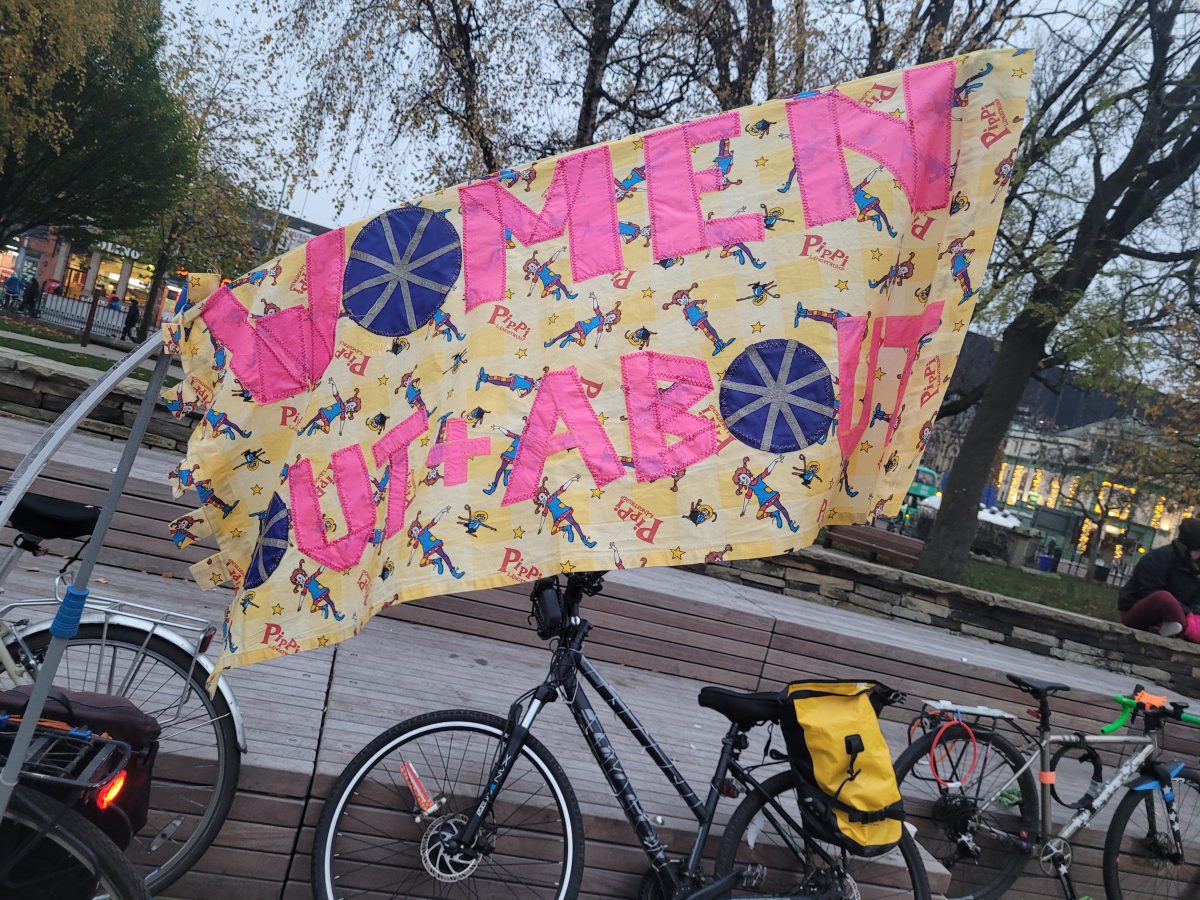 A colourful banner with text saying "Women Out + About". There are a few bicycles leaning against a wall below the banner.