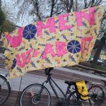 A colourful banner with text saying "Women Out + About". There are a few bicycles leaning against a wall below the banner.
