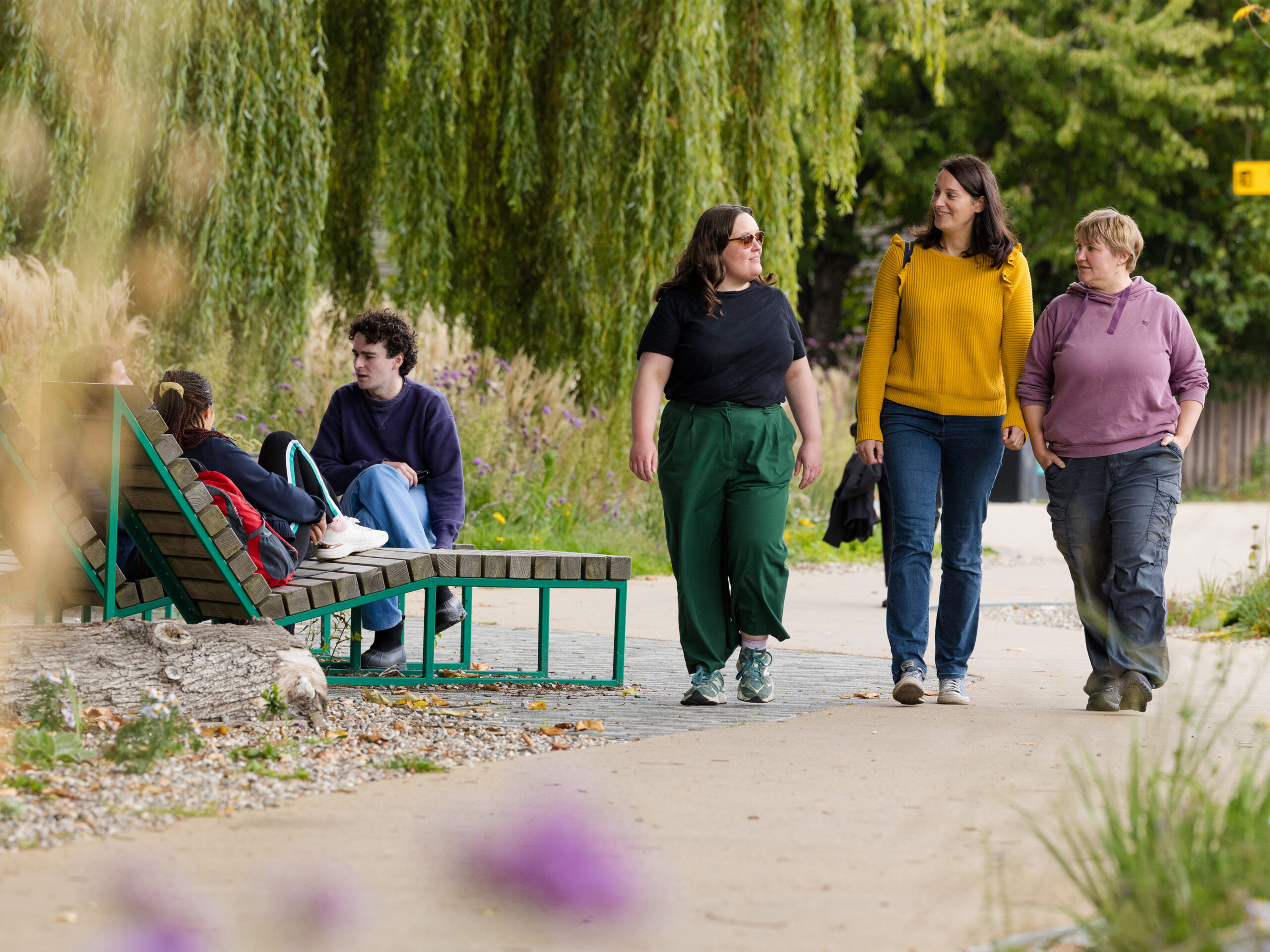 Three pedestrians walking down a street lined with SuDS and trees. Three people are sat on a sun lounger to the side. Credit: Active Travel England 2025