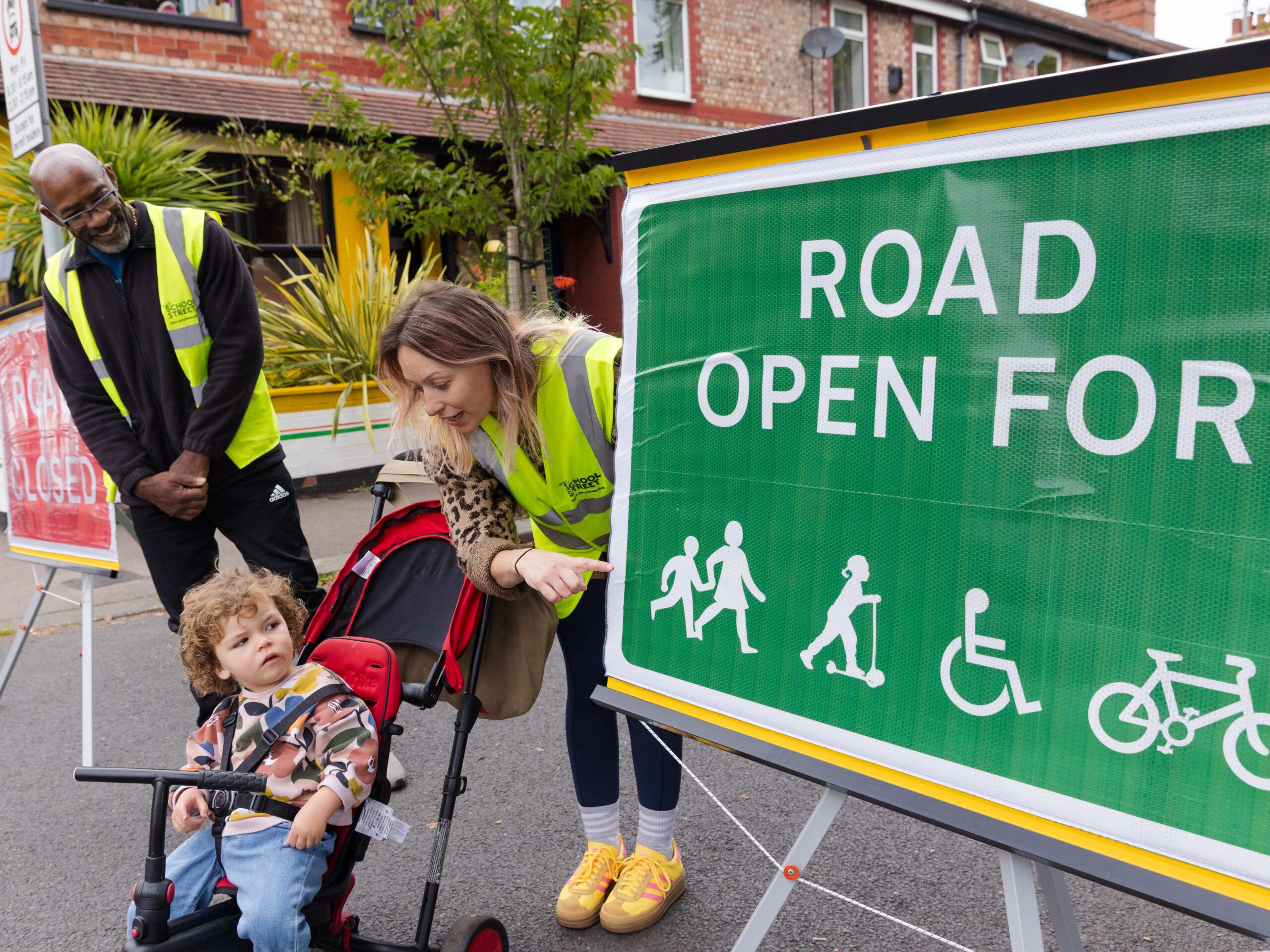 Two school streets marshals with a child in a buggy stood at the end of the road. They are stood in front of a green sign stating 'ROAD OPEN FOR' Credit: Active Travel England 2025