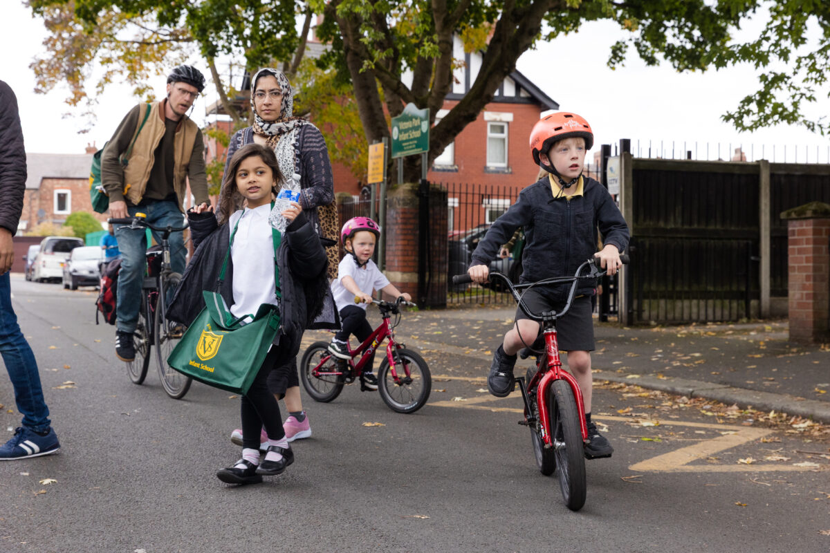 Parents and children leaving the school gate at the bell. Two children are cycling. A parent and child cross the road between them. Another parent is cycling in the background.